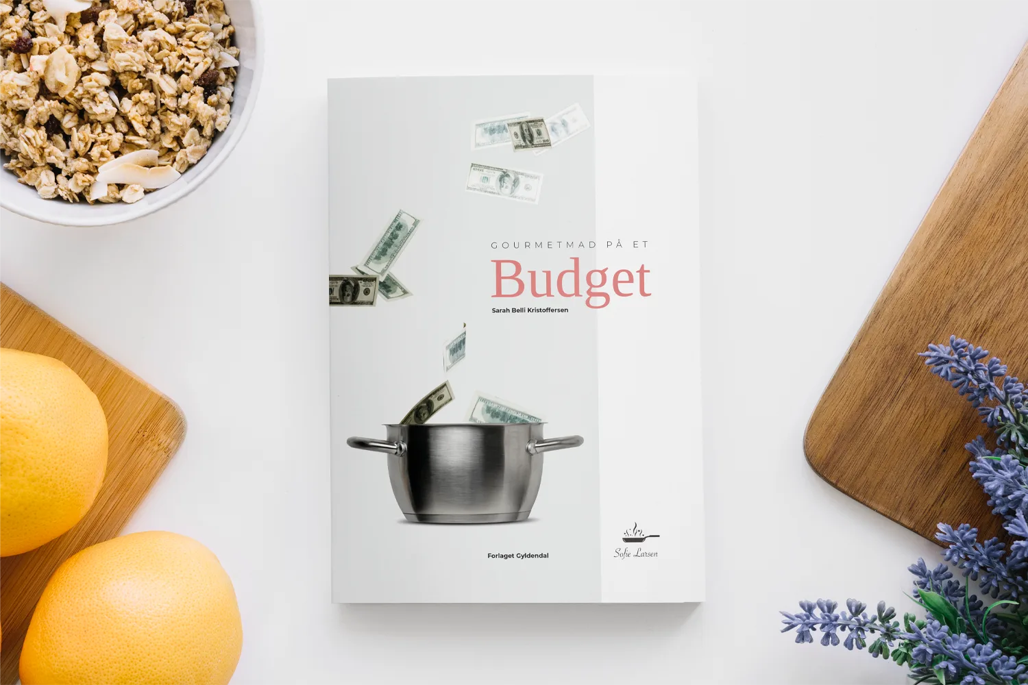 Flat lay of the book “Gourmetmad på et Budget” by Sofie Larsen, surrounded by granola, grapefruit, lavender, and a wooden board.