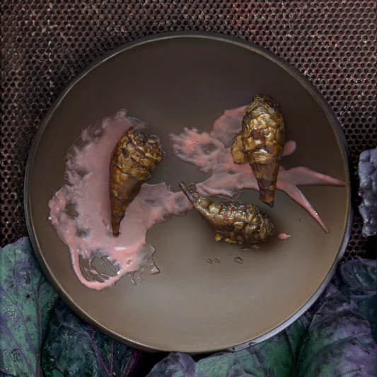 Three brownish food items on a dark plate with pink sauce, placed on a textured surface with red cabbage leaves.
