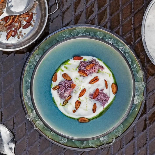 Artistic dish served in a blue bowl, featuring creamy white sauce, purple elements, and roasted nuts, surrounded by crushed ice.