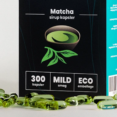 Close-up of the Elixsir Matcha syrup capsules box, featuring a green tea illustration and the text “300 kapsler,” “MILD smag,” and “ECO emballage.” Several green capsules are scattered in front of the box.