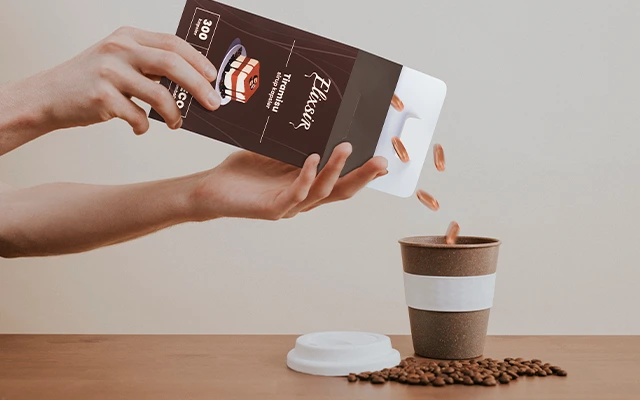 Hand pouring Tiramisu-flavored Elixsir syrup capsules from a box into a reusable coffee cup. Coffee beans are scattered on the wooden table, and the cup lid rests beside it.