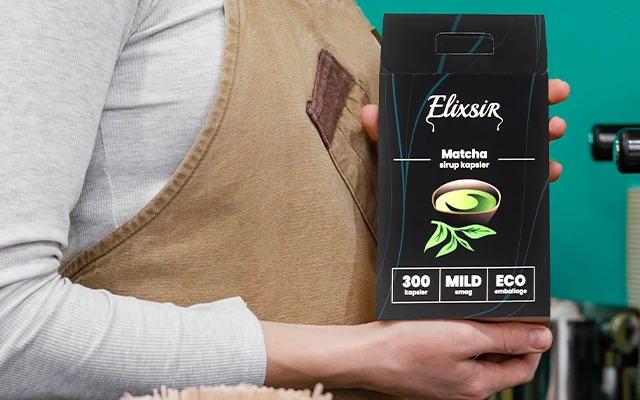 Person wearing a beige apron holding a black box of Elixsir Matcha syrup capsules. The packaging features a matcha leaf illustration and labels that read "300 capsules," "MILD taste," and "ECO packaging."