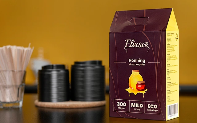 Box of Elixsir Honey syrup capsules placed on a dark counter with black coffee cups and wooden stirrers in the background. The packaging features a honey jar illustration and labels reading "300 capsules," "MILD taste," and "ECO packaging."