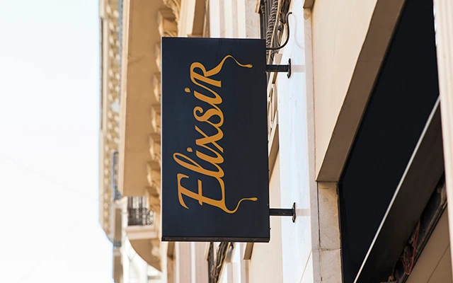 Black storefront sign with the Elixsir logo in gold lettering, mounted on the side of a building in a city street.