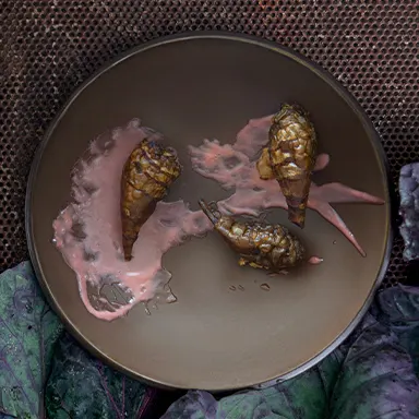 A brown ceramic plate with three artichoke-like vegetables served on a smear of pink sauce. Background includes purple cabbage leaves and a textured surface.