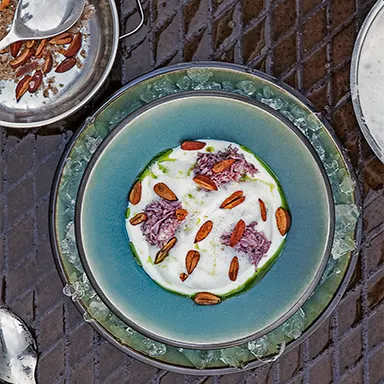 Gourmet dish with a creamy white sauce, shredded purple topping, almonds, and green oil, served in a turquoise bowl on ice.