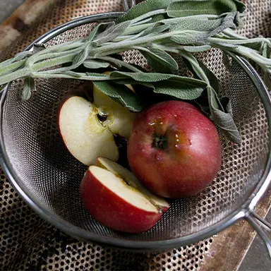 Fresh sage and sliced red apples placed in a metal strainer on a rustic surface.