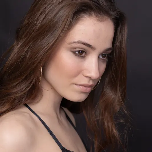 Portrait of a woman with long brown hair and subtle makeup, wearing a black strap top and gold hoop earrings. Studio-lit with a black background.