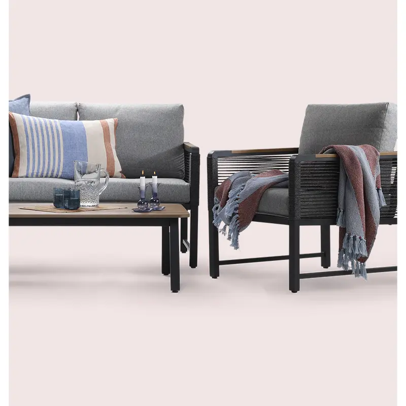 Styled product shot of grey outdoor sofa and armchair with soft cushions and blankets, placed on a light pink background.